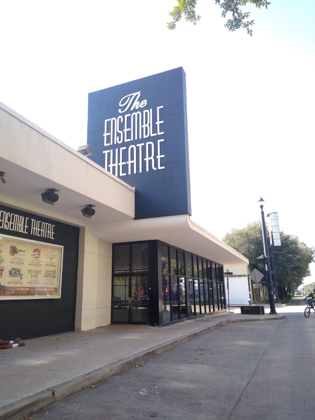 Außenansicht des Ensemble Theaters in San Francisco mit Glaswänden, einer sitzenden Person auf dem Gehweg, einem Fahrrad auf der Straße, einem Schild, einer Straßenlaterne, Bäumen und einem bewölkten Himmel.