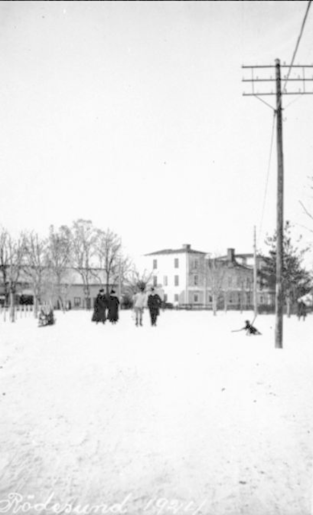Eine Gruppe von Menschen geht eine schneebedeckte Straße in Rodesund, Deutschland im Jahr 1921 entlang, gesäumt von Bäumen, Gebäuden und Strommasten, mit Text am unteren Bildrand.
