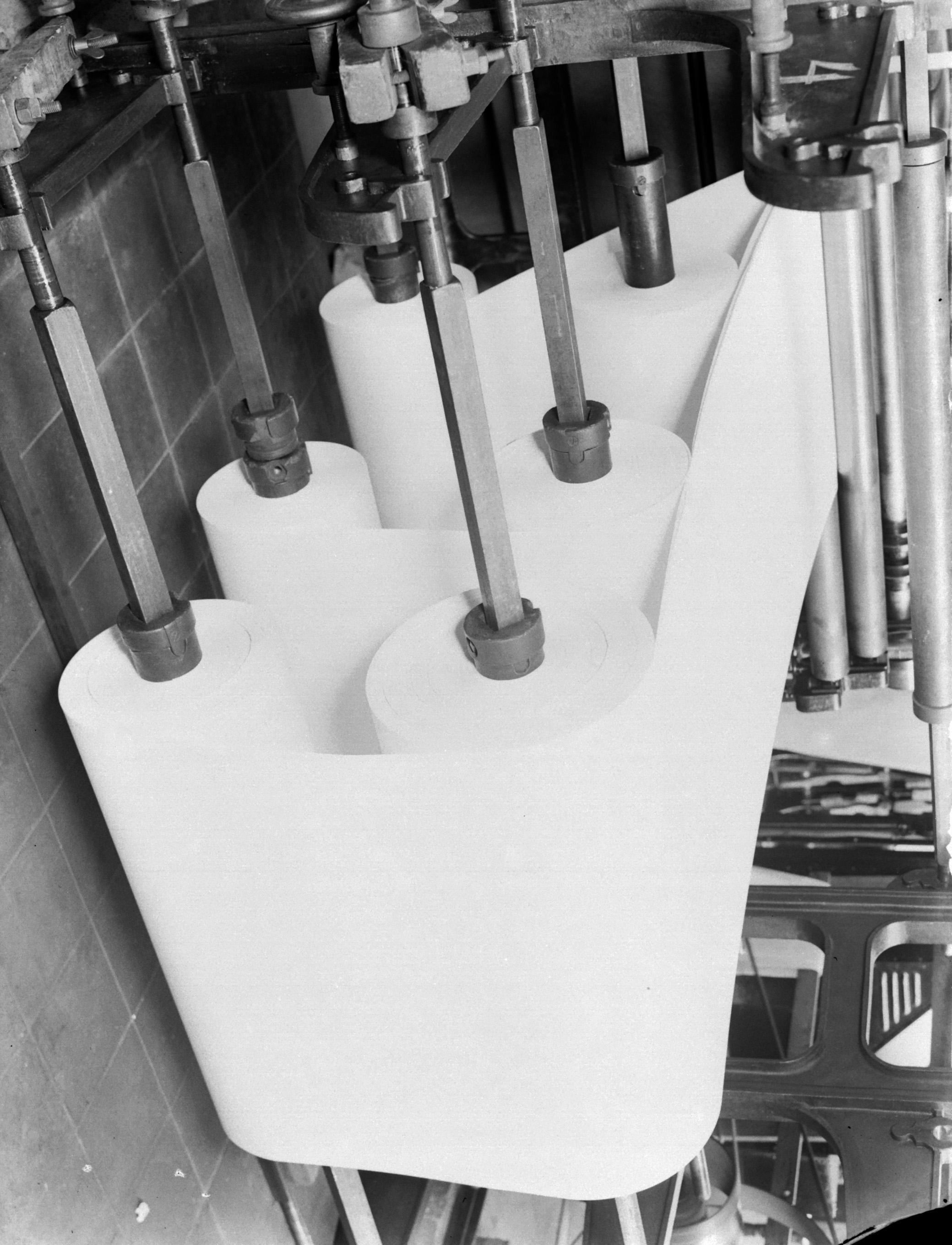 Ein Schwarz-Weiß-Foto von einem Drucker in einer Fabrik mit mehreren Papierrollen und einer Wand im Hintergrund.