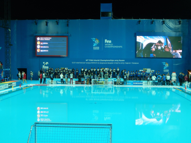 Eine Gruppe von Menschen um ein Hallenbad mit Netz am Boden herumstehend, während der FINA-Weltmeisterschaften 2015 in Kazan, mit einem Bildschirm im Hintergrund, der Text und Logos anzeigt.