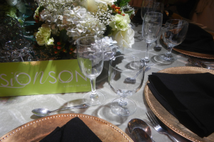 Formaler Esstisch mit goldenen Untersetzern, schwarzen Servietten, Besteck, Gläsern, einem Blumenbouquet und einer Texttafel.