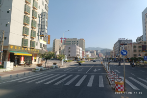 Stadtstraße mit Gebäuden, Straßenlaternen, Schildern, Fahrzeugen, Barrieren, Bäumen, Namensschildern, Strommasten, Kabel, fernen Hügeln und Himmel.
