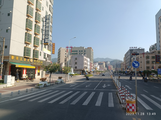 Stadtstraße mit Gebäuden, Straßenlaternen, Schildern, Fahrzeugen, Barrieren, Bäumen, Namensschildern, Strommasten, Kabel, fernen Hügeln und Himmel.