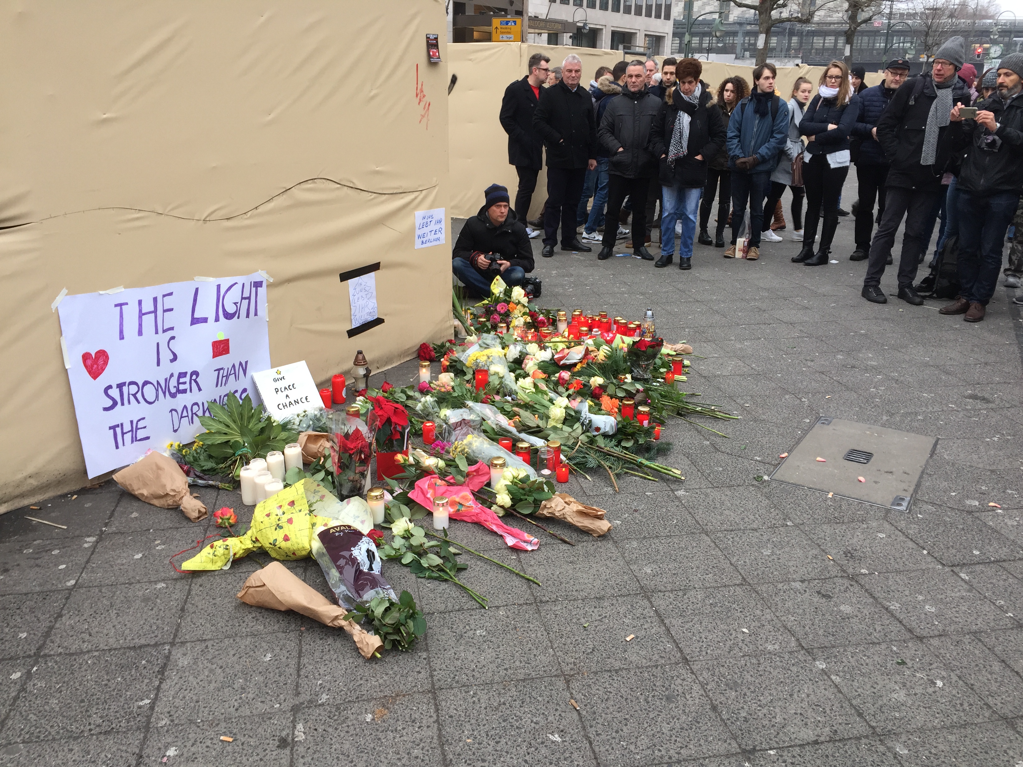 Eine Gruppe von Menschen steht vor einem Zelt mit einer Tafel, auf der "Das Licht ist stärker als die Dunkelheit" steht, umgeben von Blumensträußen, Kerzen und anderen Gegenständen, mit Gebäuden, Bäumen und einem klaren Himmel im Hintergrund.