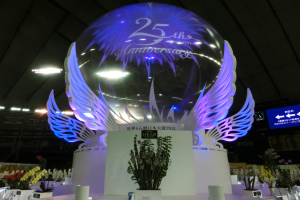 Eine große Kristallkugel mit Flügeln oben drauf, umgeben von Blumenschmuck, Namensschildern und anderen Gegenständen auf dem Boden, vor einer Kulisse aus Brettern, Lichtern und einem Bildschirm während einer 25-jährigen Jubiläumsfeier.