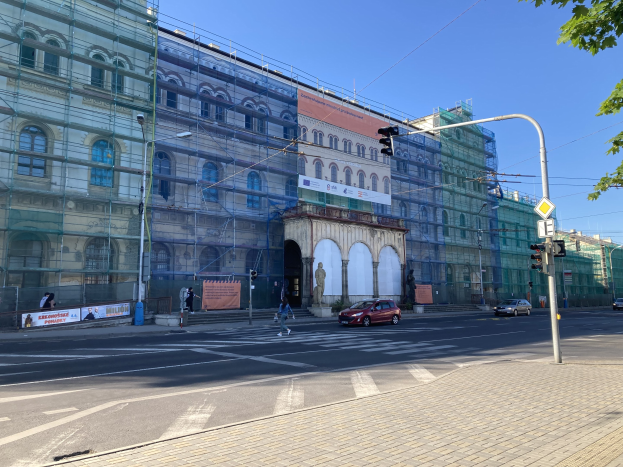 Großes Gebäude mit Gerüst während der Renovierung umgeben von Fahrzeugen, Passanten, Ampeln, Straßenschildern, Pfählen, Bäumen und einem klaren blauen Himmel.