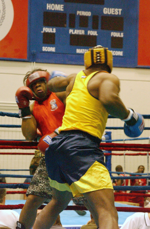 Zwei Boxer in einem Ring, umgeben von einer Menge, mit einer Tafel, die Text und Logos an der Wand dahinter anzeigt.