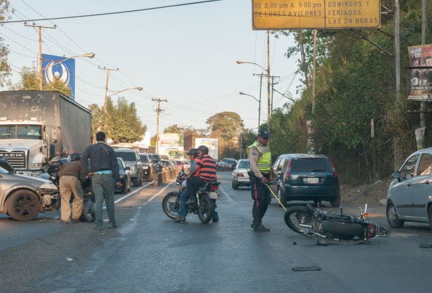 Eine Gruppe von Menschen steht um ein verunglücktes Motorrad auf der Straße mit mehreren Fahrzeugen, darunter ein Lastwagen, und Hintergrundelementen wie Bäumen, Pfählen, Lampen und Schildern.
