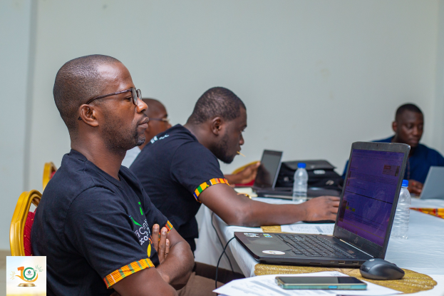 Gruppe von M├Ąnnern, die um einen Tisch sitzen und Laptops, Wasserflaschen, Mobiltelefone und Papiere haben; Wand im Hintergrund zeigt das Logo und den Text 'Nigerianische Regierung startet Digital Governance-Programm'.