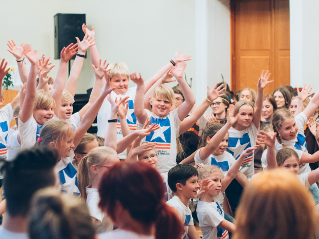 Eine Gruppe von Kindern steht mit erhobenen Händen vor einer Menge, mit einer Wand und einer Tür im Hintergrund; ein Tisch mit einer Vase und einem Lautsprecher ist auf der linken Seite.