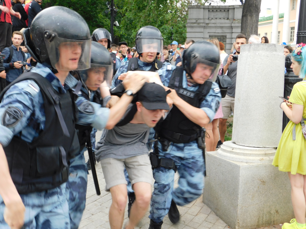Eine Gruppe von Einsatzpolizisten in Ausrüstung, die vor einer Menge davonläuft, einige halten Kameras, mit einer Säule im Vordergrund, Bäumen, Gebäuden und Laternen im Hintergrund.