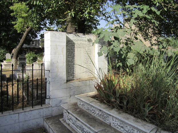 Ein Blick auf einen Friedhof mit Treppe unten, Gras und Bäumen rechts, einem Gedenkstein in der Mitte, einem Geländer daneben, Gräbern dahinter und Bäumen und Gebäuden im Hintergrund.