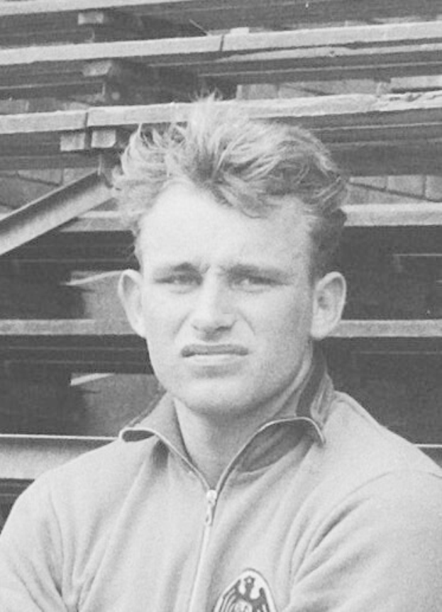 Schwarzes und weißes Foto eines Mannes in einer Jacke vor gestapelten Holzplanken, der angeblich ein legendärer deutscher Fußballspieler ist, der im Alter von 89 Jahren verstorben ist.