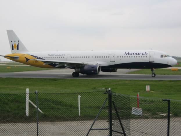 Ein Monarch Airlines Airbus A320-200 auf einer von Gras, einem Zaun mit Pfählen und Brettern umgebenen Rollbahn.