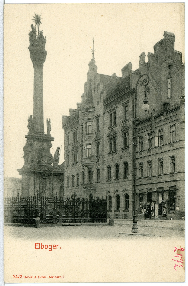 Ein altes Schwarz-Weiß-Foto eines Stadtplatzes mit einem Denkmal in der Mitte, umgeben von Gebäuden, Straßenlaternen, Gittern und einem klaren Himmel, mit Text am unteren Bildrand.