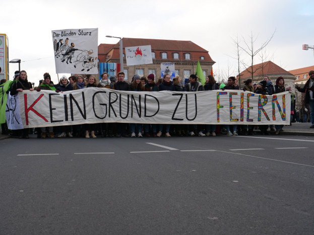 Eine Gruppe von Menschen steht auf einer Straße und hält ein Banner mit der Aufschrift "Kein Grund zu Feiern" im Protest, mit Gebäuden, Laternenmasten, Bäumen und einem klaren blauen Himmel im Hintergrund.