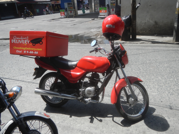 Zwei Motorr√°der nebeneinander auf einer Straße geparkt, eines mit roter Box und Helm darauf, mit Gebäuden, Geschäften, Schildern, Masten und anderen Fahrzeugen im Hintergrund.