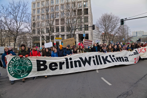 Eine Gruppe von Menschen geht eine von Bäumen gesäumte Straße entlang und hält ein Banner mit der Aufschrift "Berlin Willklima" hoch, mit Gebäuden und einer Ampel im Hintergrund unter einem bewölkten Himmel.