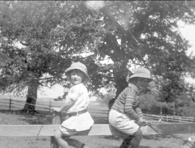 Zwei junge Jungs in Hüten mit Baseballschlägern in einem Park mit Bäumen, einem Zaun und Gras, in Schwarz-Weiß.