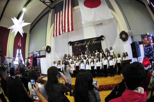 Eine Gruppe von Menschen auf einer Bühne vor einem Publikum, einige halten Kameras und Telefone, mit geschmückten Weihnachtsbäumen, einem Podium mit Mikrofon, Lautsprechern, einer Texttafel, Vorhängen, Flaggen und Deckenleuchten.