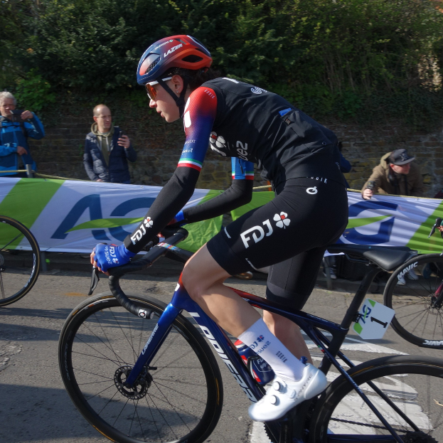 Professionelle Radsportlerin Katie Archibald mit Helm, Schutzbrille, Handschuhen und Schuhen fährt auf einem Fahrrad eine Straße entlang neben einer Menge, mit einem Banner, einer Wand, Bäumen und einem Gebäude im Hintergrund.