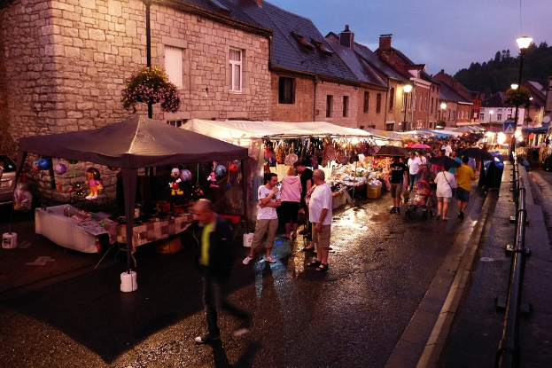 Eine Gruppe von Menschen auf einer Straße mit Geschäften, Schirmen, hängenden Spielzeugen, Taschen, Gebäuden mit Fenstern, Bäumen, Straßenlaternen und Absperrungen.