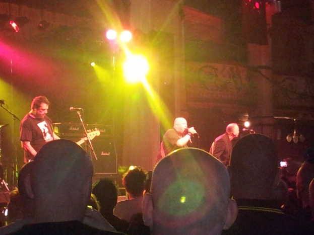 Drei Männer auf der Bühne bei einem Konzert, einer spielt Gitarre, eine große Menge davor und Lichter und ein Gebäude im Hintergrund.