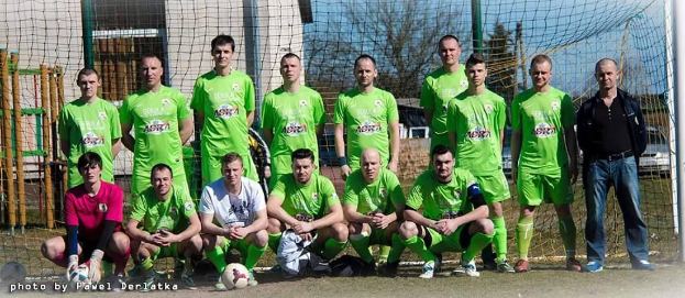 Gruppe von Männern in grünen T-Shirts auf einem Fußballfeld mit Ball und Tor, Bäumen und Gebäuden im Hintergrund.