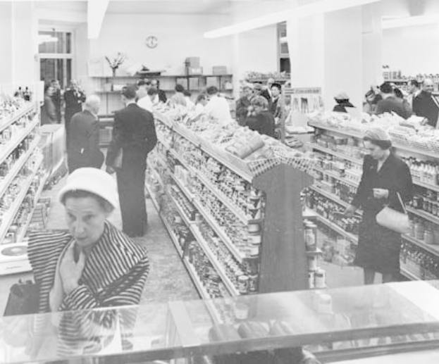 Schwarz-weiß-Foto von Menschen beim Einkaufen in einem Supermarkt mit Regalen voller Lebensmittel, einige halten Taschen, eine Uhr an der Wand und Deckenbeleuchtung.