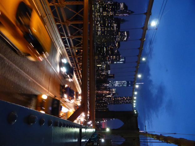 Ein gelbes Taxi fährt nachts über die beleuchtete Brooklyn Bridge, mit Gebäuden und Sternen im Hintergrund.