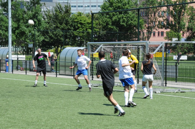 Personen in Kleidung beim Fuballspielen auf einem Feld mit einem Zaun und einem Tor in der Mitte, einem Pfosten und einem Fuball oben links und Bäumen und Gebäuden im Hintergrund.