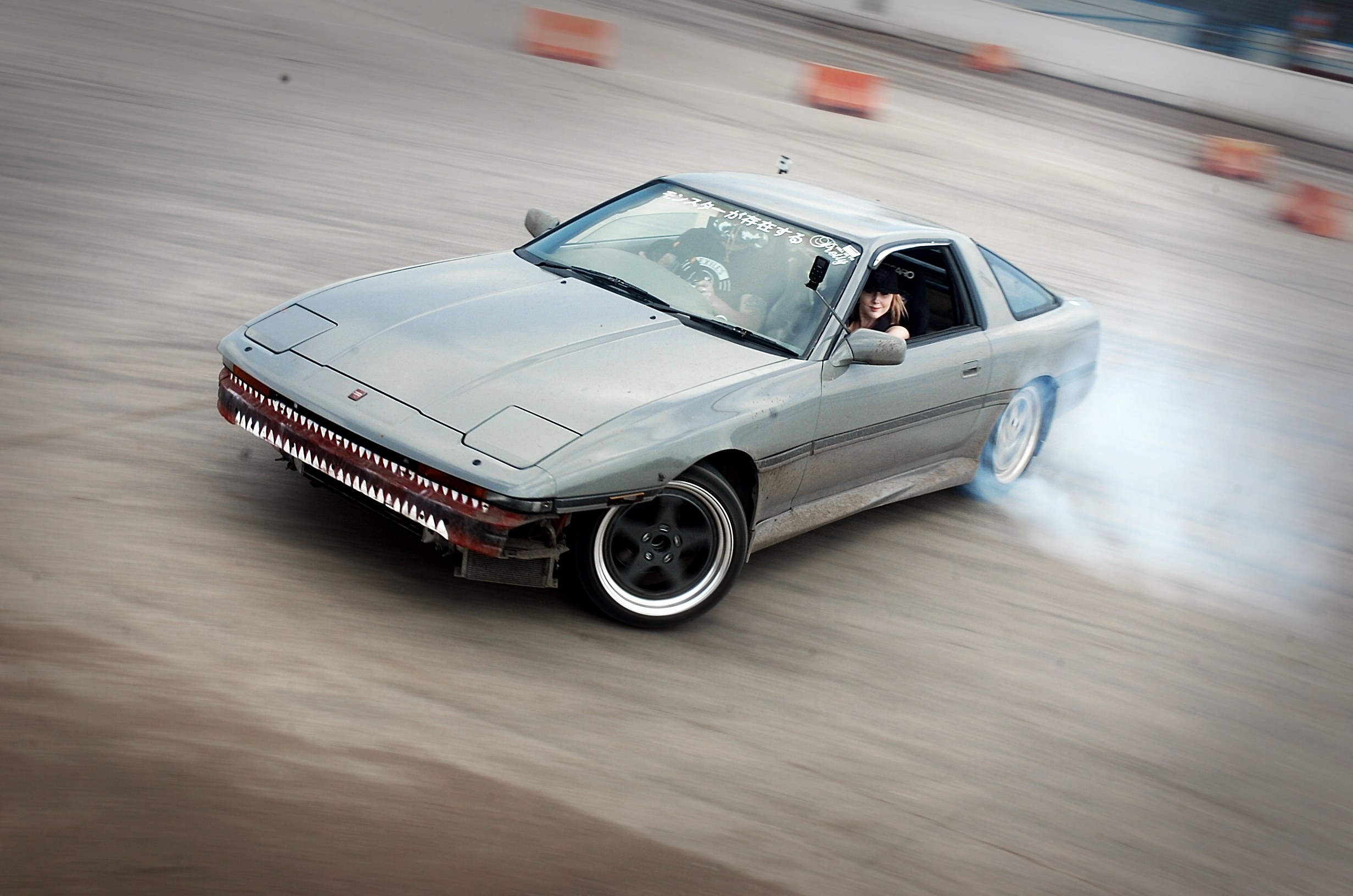 Eine Person in einem Helm fährt ein Auto auf einer Rennstrecke, mit Rauch, umgeben von orangen Objekten, mit einem unscharfen Hintergrund, der hohe Geschwindigkeit andeutet.