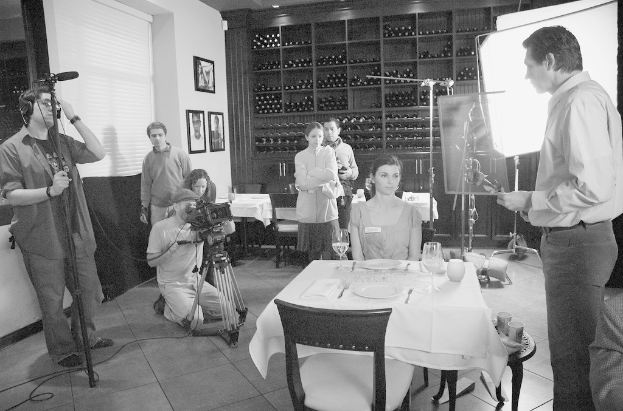 Eine Gruppe von Menschen auf einem Filmset, mit einem Mann auf der rechten Seite, einer Frau an einem Tisch mit Tellern, Gabeln und Gl√§sern in der Mitte und zwei Personen, die eine Kamera bedienen, auf der linken Seite, mit Regalen, Rahmen und einem Bildschirm im Hintergrund.