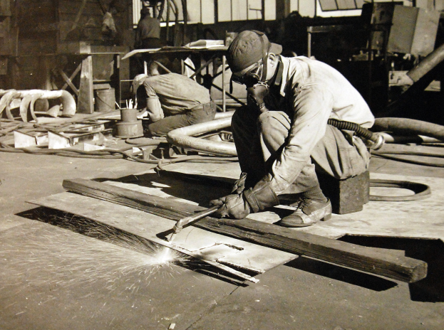 Ein Schweißer in einem Helm, Schutzbrille und Handschuhen arbeitet an einem Holzstück in einer Fabrik und hält ein Schweißwerkzeug in der Hand, mit Rohren, Stäben und anderen Gegenständen im Hintergrund.