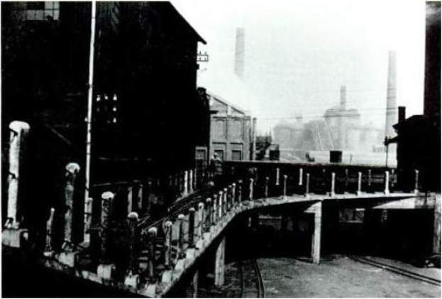 Ein Schwarz-Wei\ss-Foto einer Br"cke "uber eine Bahnschiene, mit Geb"uden auf beiden Seiten und einer Fabrik im Hintergrund, mit Gel"ndern und Pf"hlen entlang der Seiten der "Uberbr"cke.