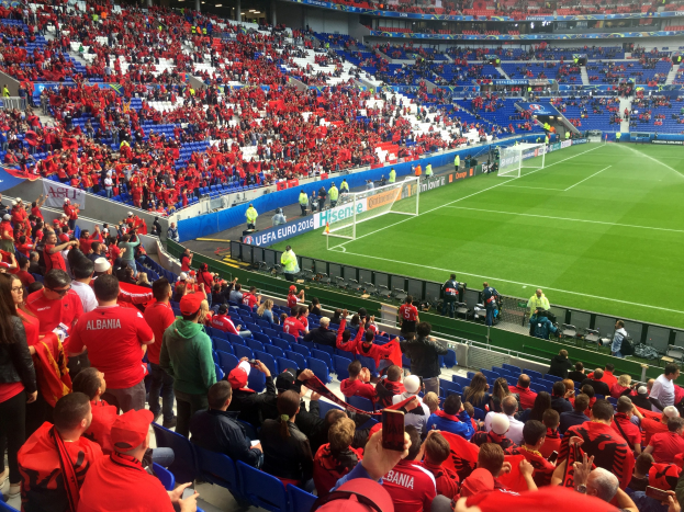 Eine große Menge Menschen in einem Stadion schaut einem Fussballspiel zu, mit Menschen auf Sitzen und Stehplätzen, Bannern, einem Torpfosten und Deckenlichtern.