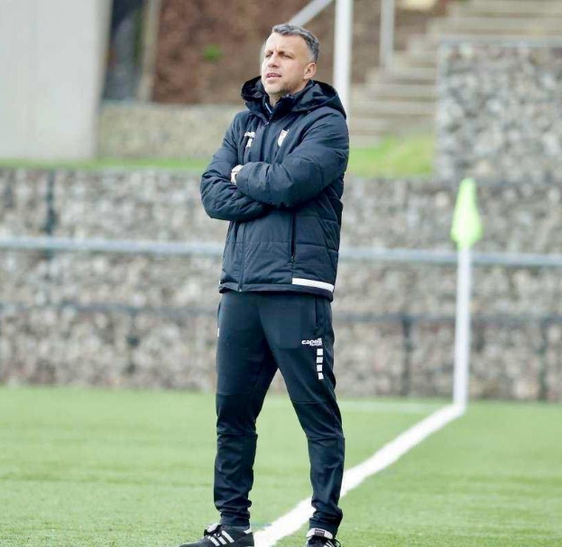 Ein Mann in einer schwarzen Jacke und Hose steht auf einem Fussballfeld mit überkreuzten Armen, der als Trainer während einer Trainingseinheit erscheint, mit einem Zaun, Treppe und Wand im Hintergrund.