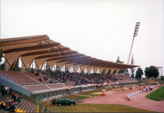 Altes Schwarz-Weiß-Foto eines Stadions voller Zuschauer, das sitzende Fans unter dem Dach, ein geparktes Auto, stehende Zuschauer auf dem Feld, Stützstangen, einen Metallrahmen, einen Zaun, eine Straßenlaterne, Bäume, einen fernen Turm und einen bewölkten Himmel zeigt.
