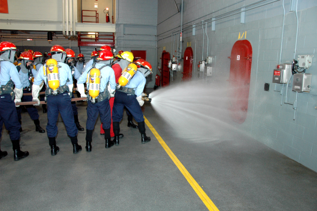 Eine Gruppe von Feuerwehrleuten in Helmen und Zylindern sprüht sich gegenseitig mit Wasser in einem Raum mit Rohren, Feuerlöschern, einer Leiter und anderen Gegenständen an den Wänden.