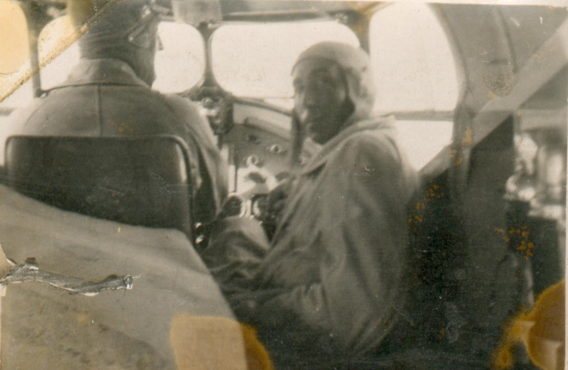 Schwarzes und weißes Foto von zwei Männern in einem Flugzeugcockpit, einer sitzt im Pilotensitz und trägt eine Uniform, der andere steht neben ihm, beide haben ernste Gesichter.