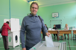 Ein Mann steht vor einer Wahlkabine und hält ein Blatt Papier in der Hand, mit Menschen, die hinter ihm sitzen und stehen, und einem Basketballkorb an der Wand im Hintergrund.
