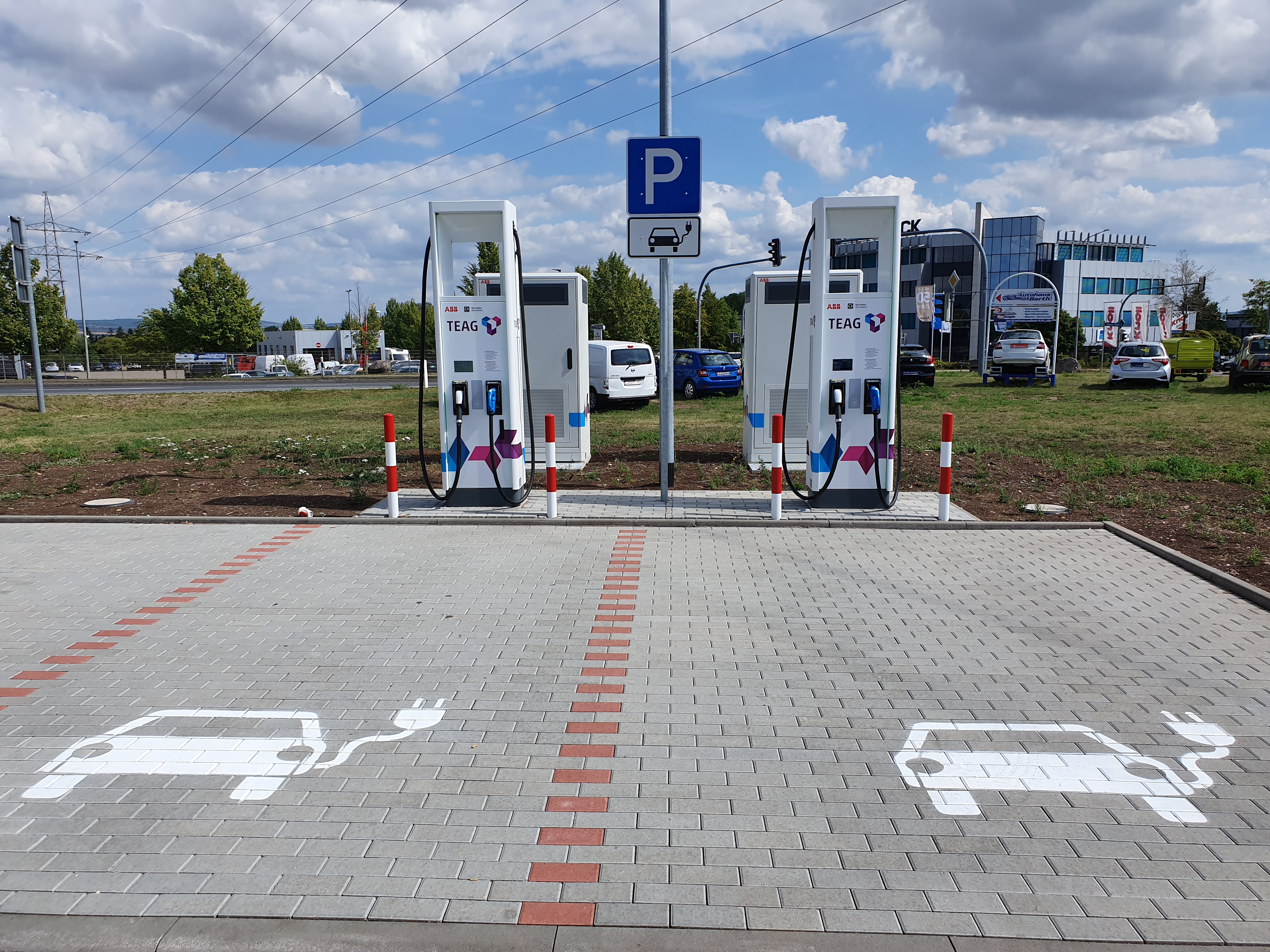 Eine Elektroauto-Ladestation auf einem Parkplatz umgeben von Gebäuden, Bäumen, Strommasten mit Drähten und Fahrzeugen auf der Straße, mit bewölktem Himmel und grasbewachsenem Boden.
