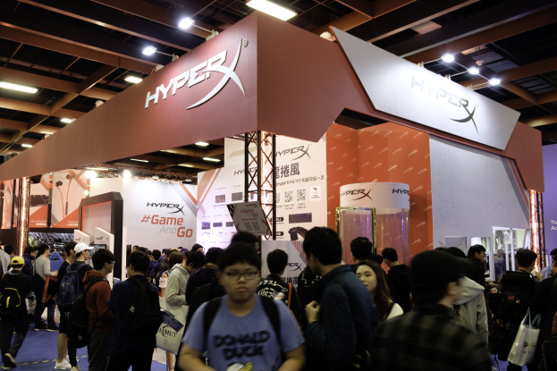 Eine große Menschenmenge versammelt sich vor einem Gaming-Expo-Stand, wobei Besucher Taschen und Kappen tragen, die von Deckenleuchten beleuchtet werden.