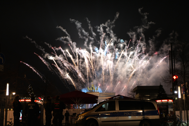 Ein Polizeiwagen steht vor einer Menschenmenge, im Hintergrund sind Geb├Ąude, B├Ąume, Masten, Verkehrszeichen und Schilder beleuchtet, w├Ąhrend farbige Feuerwerke den Himmel erhellen, w├Ąhrend des Silvesterfeuerwerks in Berlin.