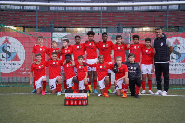 Eine Gruppe junger Männer in roten T-Shirts und schwarzen Shorts steht auf einem Fußballfeld neben einer Kiste roter Gegenstände, im Hintergrund sind Banner mit "FC Rot-Schwarz Eglaute - Champions League" zu sehen.