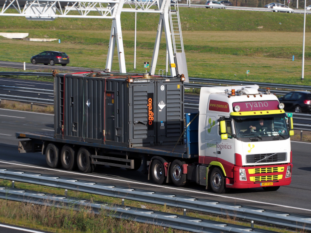 Ein Lastwagen mit einem großen Behälter fährt auf einer Autobahn, mit anderen Fahrzeugen, Masten, Bäumen, Gebäuden und einem klaren blauen Himmel im Hintergrund.