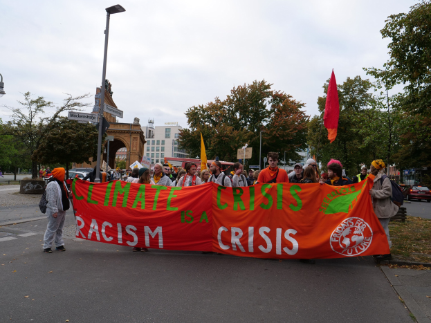 Eine Gruppe von Menschen marschiert eine baumbestandene Straße entlang und hält ein Banner mit der Aufschrift "Klimakrise ist eine Krise", während Autos vorbeifahren und Gebäude im Hintergrund zu sehen sind, unter einem klaren blauen Himmel.