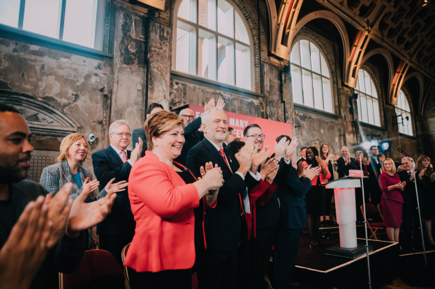 Eine Gruppe von Menschen klatscht feierlich vor einem Publikum, mit einem Podium mit Mikrofon auf der rechten Seite und einem Banner im Hintergrund, das "Covid-19 New York City Mayor Bill de Blasio" lautet.