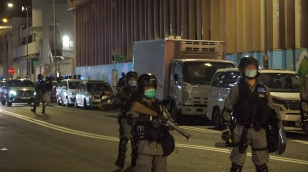 Polizeibeamte in Helmen, Handschuhen und Gesichtsmasken stehen auf einer Straße in der Nacht, einige halten Waffen, mit Fahrzeugen, Laternenmasten, Schildern und Gebäuden im Hintergrund.
