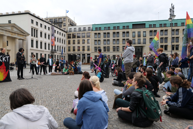 Eine Gruppe von Menschen, die auf dem Boden vor einer Menge sitzt, die Fahnen und Transparente mit Text hält, mit einer Person, die in ein Mikrofon spricht, einer Statue auf einem Sockel und Gebäuden im Hintergrund während einer Anti-Schwulen-Demo in Berlin.